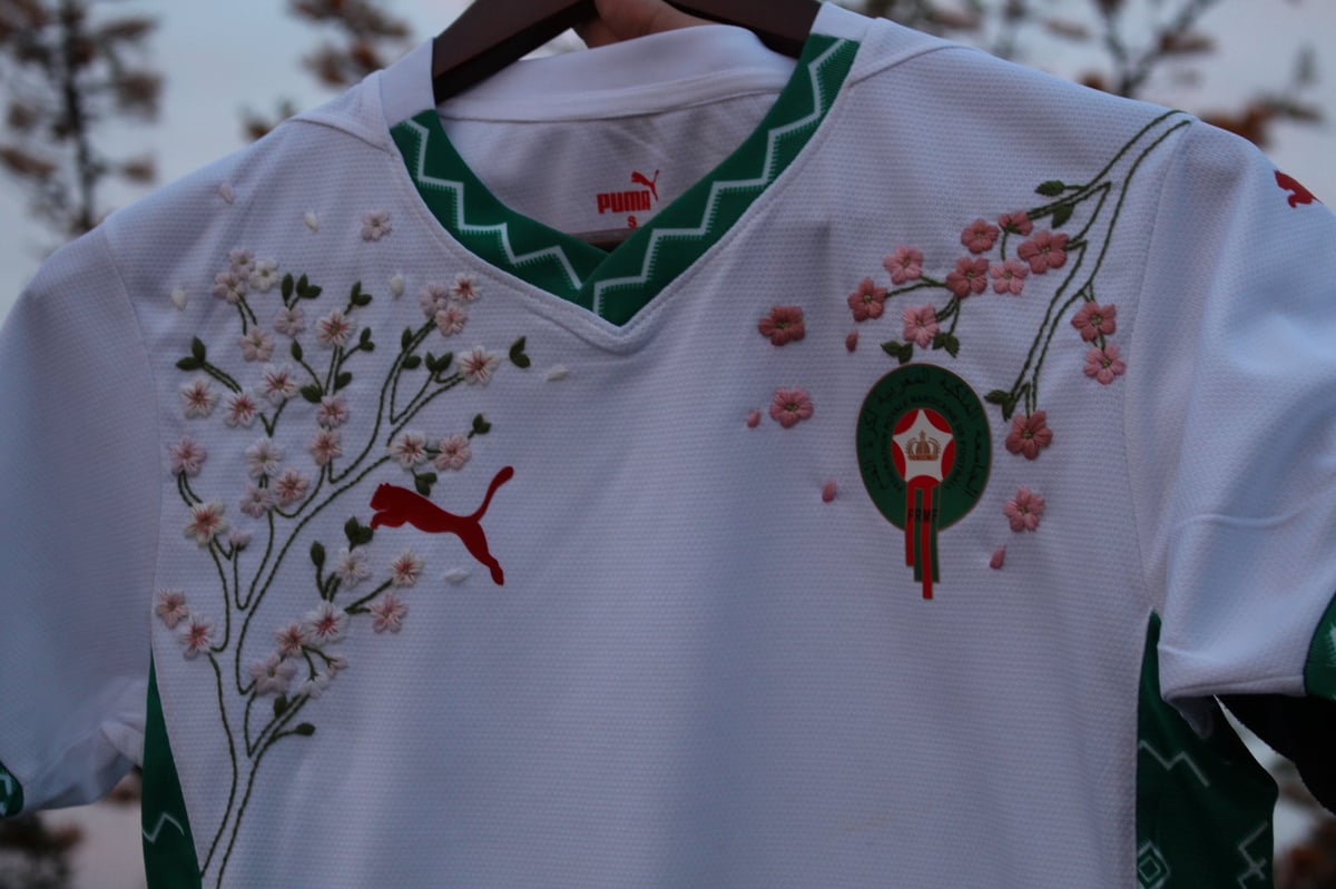 White tunic with green trim and embroidered flowers and bird design, hanging on wooden hanger outdoors
